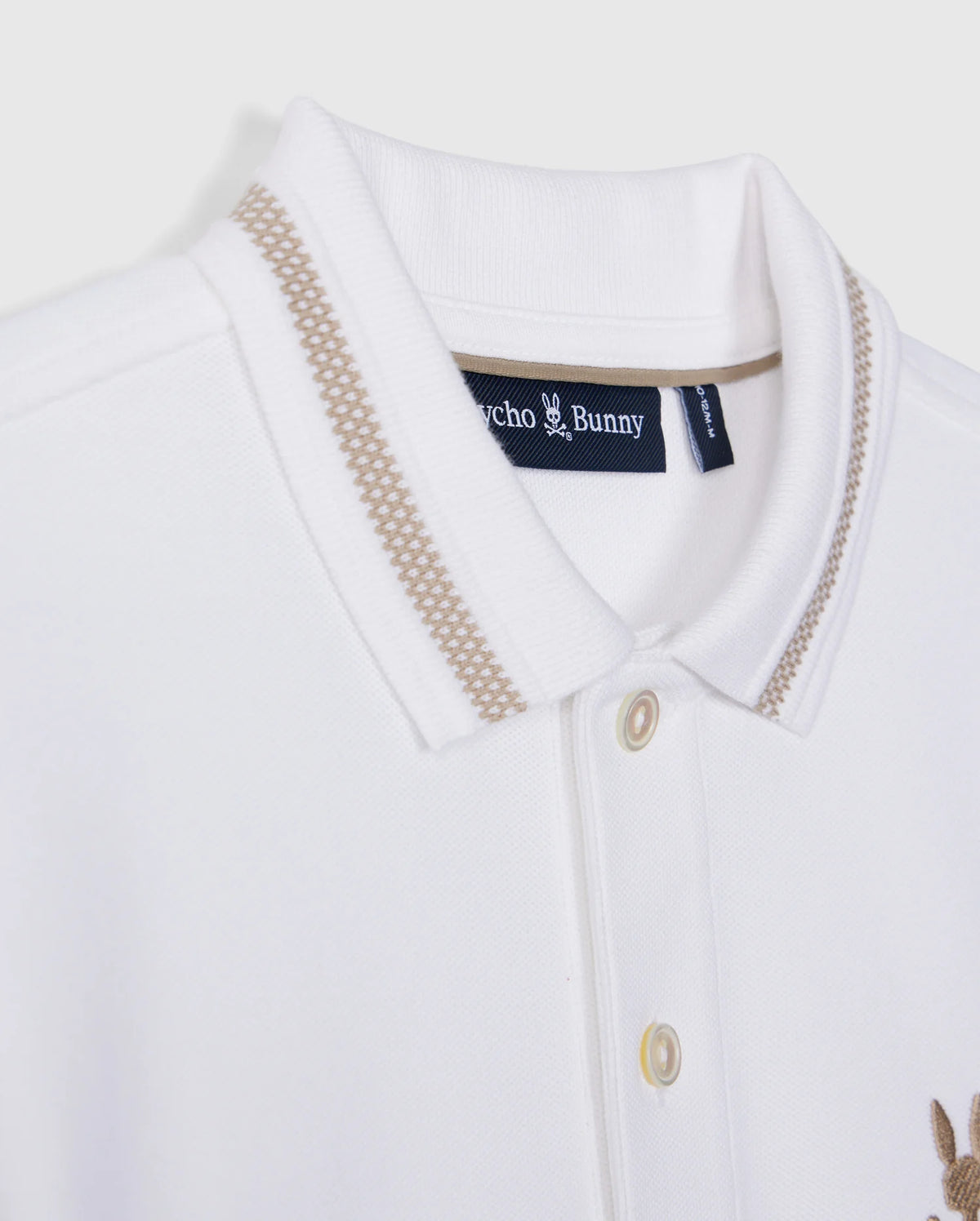 Close-up of the Kids Riviera Pique Polo Shirt - B0K640C200 from Psycho Bunny, a white polo featuring beige accents on the collar. It includes a small "Psycho Bunny" tag on the inside of the collar and is crafted from soft Pima cotton. The shirt has a button-front placket and an embroidered, subtle beige bunny logo on the chest.