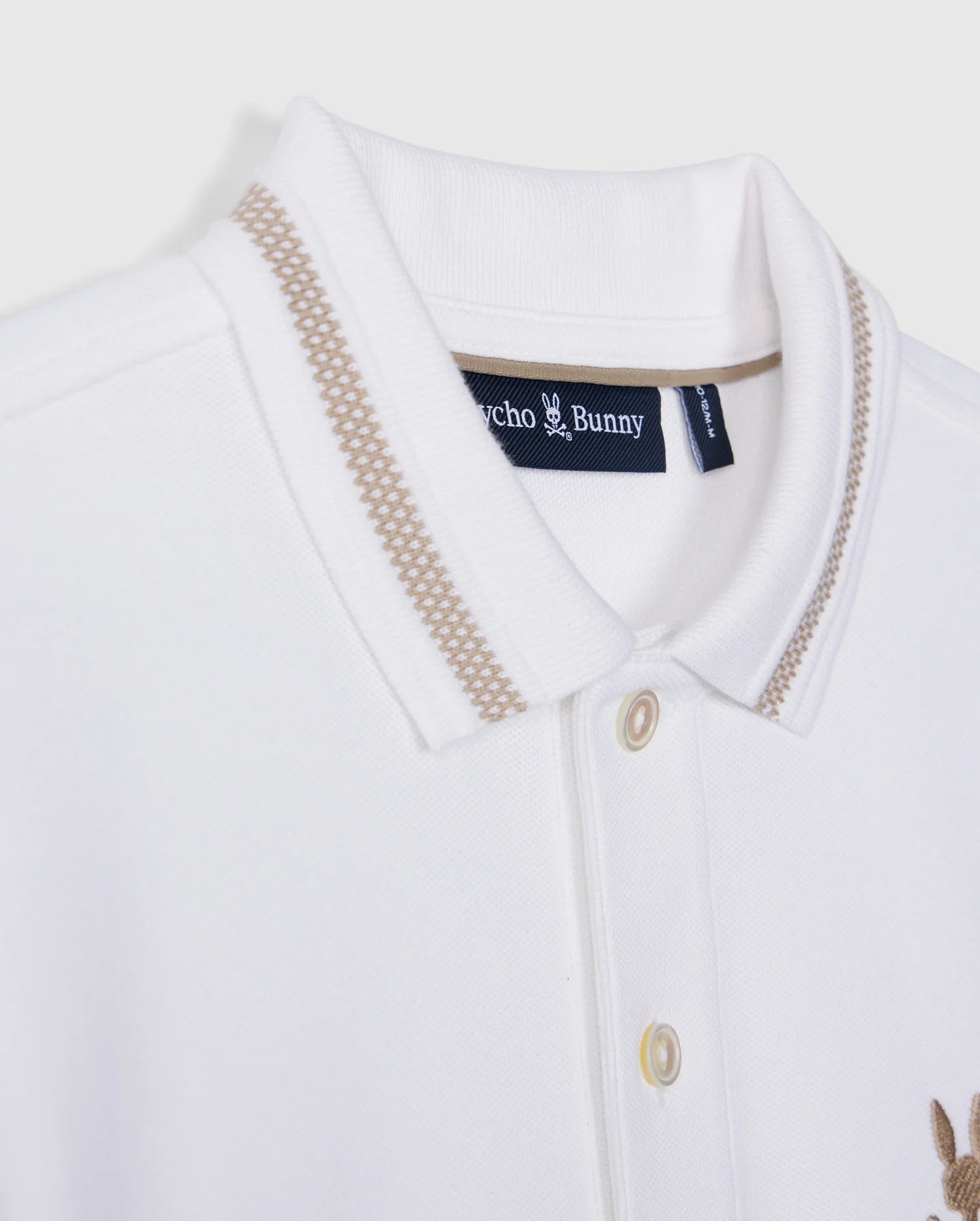 Close-up of the Kids Riviera Pique Polo Shirt - B0K640C200 from Psycho Bunny, a white polo featuring beige accents on the collar. It includes a small "Psycho Bunny" tag on the inside of the collar and is crafted from soft Pima cotton. The shirt has a button-front placket and an embroidered, subtle beige bunny logo on the chest.