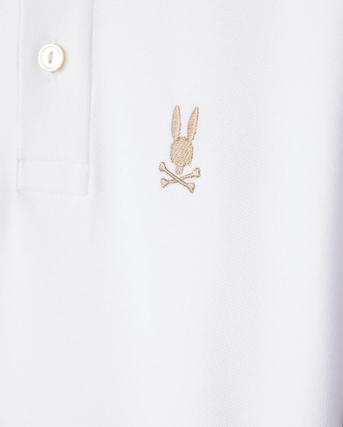 Here's a close-up of the Psycho Bunny KIDS RIVIERA PIQUE POLO SHIRT - B0K640C200 in white, crafted from soft Pima cotton. The shirt features an embroidered chest Bunny logo consisting of a light brown rabbit head above crossed bones. It has a standard collar with a button placket and one visible button.