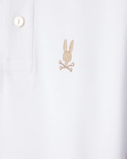 Here's a close-up of the Psycho Bunny KIDS RIVIERA PIQUE POLO SHIRT - B0K640C200 in white, crafted from soft Pima cotton. The shirt features an embroidered chest Bunny logo consisting of a light brown rabbit head above crossed bones. It has a standard collar with a button placket and one visible button.