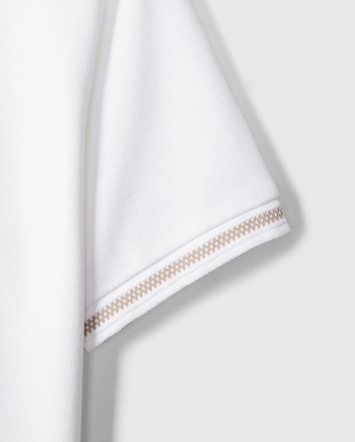 Close-up of the sleeve of a Kids Riviera Pique Polo Shirt - B0K640C200 by Psycho Bunny, made from textured Pima cotton fabric. The hem of the sleeve features two rows of intricate beige stitching, adding a decorative element to the minimalist design. The background is plain and neutral, ensuring the focus remains on the sleeve.
