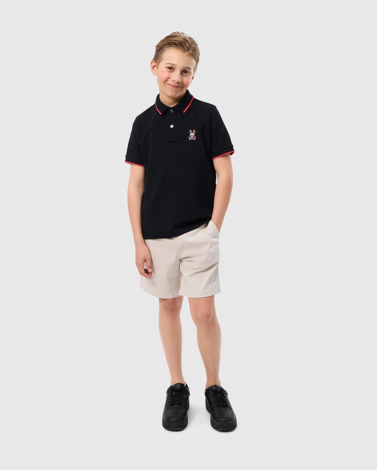 A young boy stands against a plain background, smiling slightly. He is wearing a Psycho Bunny KIDS KAYDEN PIQUE POLO - B0K675C200 with bold contrasting stripes, beige shorts, and black shoes. One hand is in his pocket while the other rests by his side. He has short, light brown hair.
