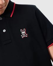Close-up of a person wearing the Psycho Bunny KIDS KAYDEN PIQUE POLO (B0K675C200) in black, featuring bold contrasting stripes and red trim. Crafted from Pima cotton piqué, this polo shirt showcases an embroidered logo of a skull with bunny ears and crossbones on the left chest. Only part of the person's face and torso are visible.