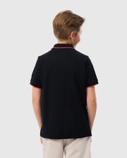 A young boy stands with his back to the camera, wearing a black KIDS KAYDEN PIQUE POLO - B0K675C200 from Psycho Bunny, featuring red trim on the collar and sleeves. Made from Pima cotton piqué, it complements his light brown hair and beige pants against a plain white background.