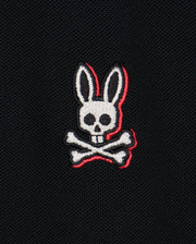 A logo featuring a white skull with rabbit ears and two crossed bones beneath it, embroidered on a black Pima cotton piqué fabric background. The skull and bones have red outline accents, adding striking detail to the bold contrasting stripes of the Psycho Bunny Kids Kayden Pique Polo (B0K675C200).