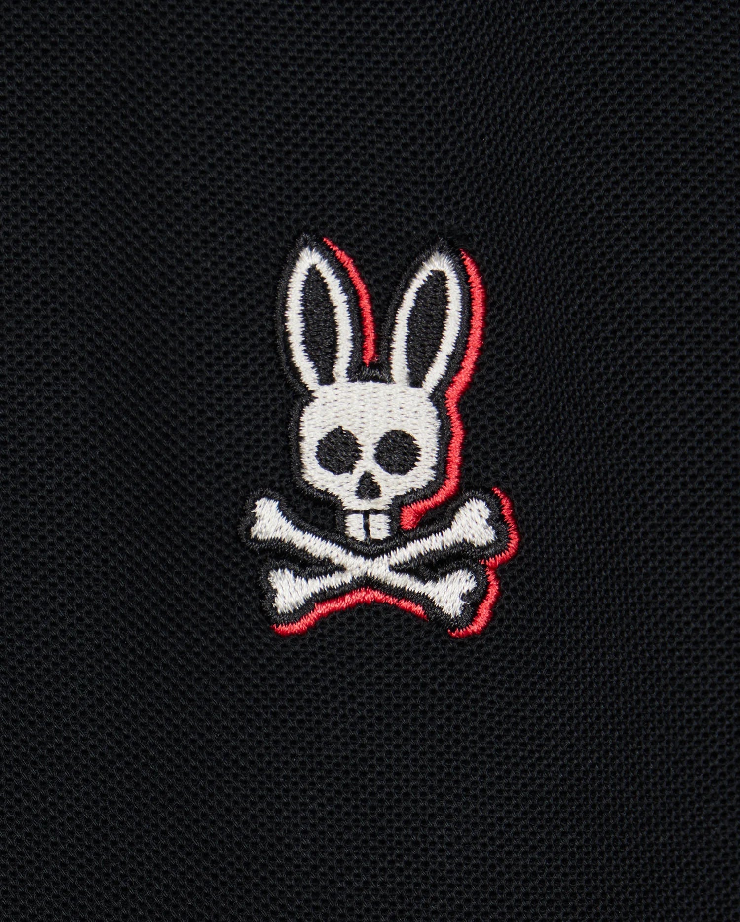 A logo featuring a white skull with rabbit ears and two crossed bones beneath it, embroidered on a black Pima cotton piqué fabric background. The skull and bones have red outline accents, adding striking detail to the bold contrasting stripes of the Psycho Bunny Kids Kayden Pique Polo (B0K675C200).