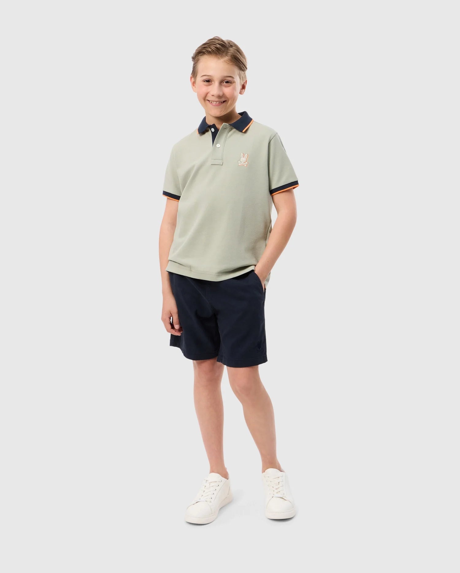 A young boy stands smiling with one hand in his pocket. He is wearing a light-colored Kids Kayden Pique Polo - B0K675C200 by Psycho Bunny, which features a small bunny logo on the chest, navy blue shorts, and white sneakers. The background is plain and white.