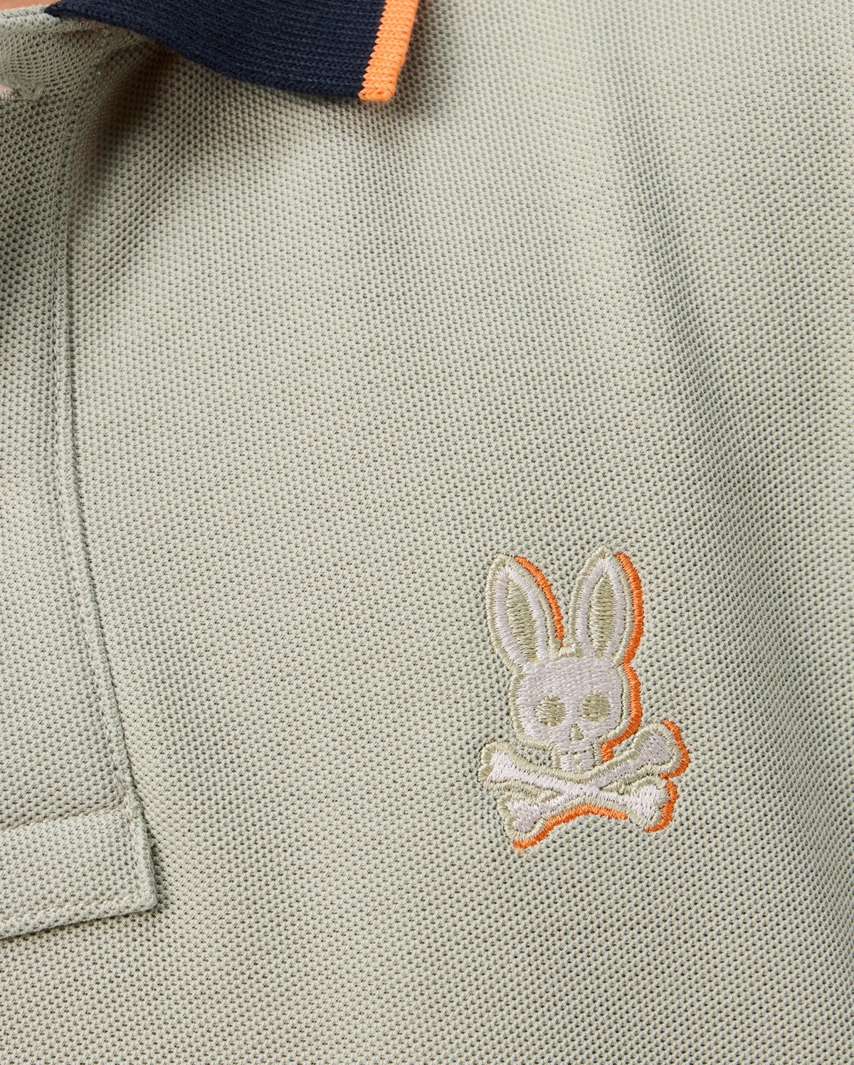 Close-up of the Psycho Bunny KIDS KAYDEN PIQUE POLO - B0K675C200, a light gray Pima cotton pique polo shirt with a dark blue collar and orange trim. The shirt is adorned with a small, embroidered bunny skull and crossbones emblem on the chest in white thread with an orange outline.