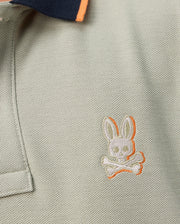 Close-up of the Psycho Bunny KIDS KAYDEN PIQUE POLO - B0K675C200, a light gray Pima cotton pique polo shirt with a dark blue collar and orange trim. The shirt is adorned with a small, embroidered bunny skull and crossbones emblem on the chest in white thread with an orange outline.