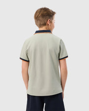 A young boy with short, light brown hair is seen from the back, wearing a Psycho Bunny KIDS KAYDEN PIQUE POLO - B0K675C200 in light gray with dark navy collar and sleeve trims, featuring contrasting stripes. The Pima cotton pique shirt pairs perfectly with his dark navy shorts as he stands against a plain, light gray background.