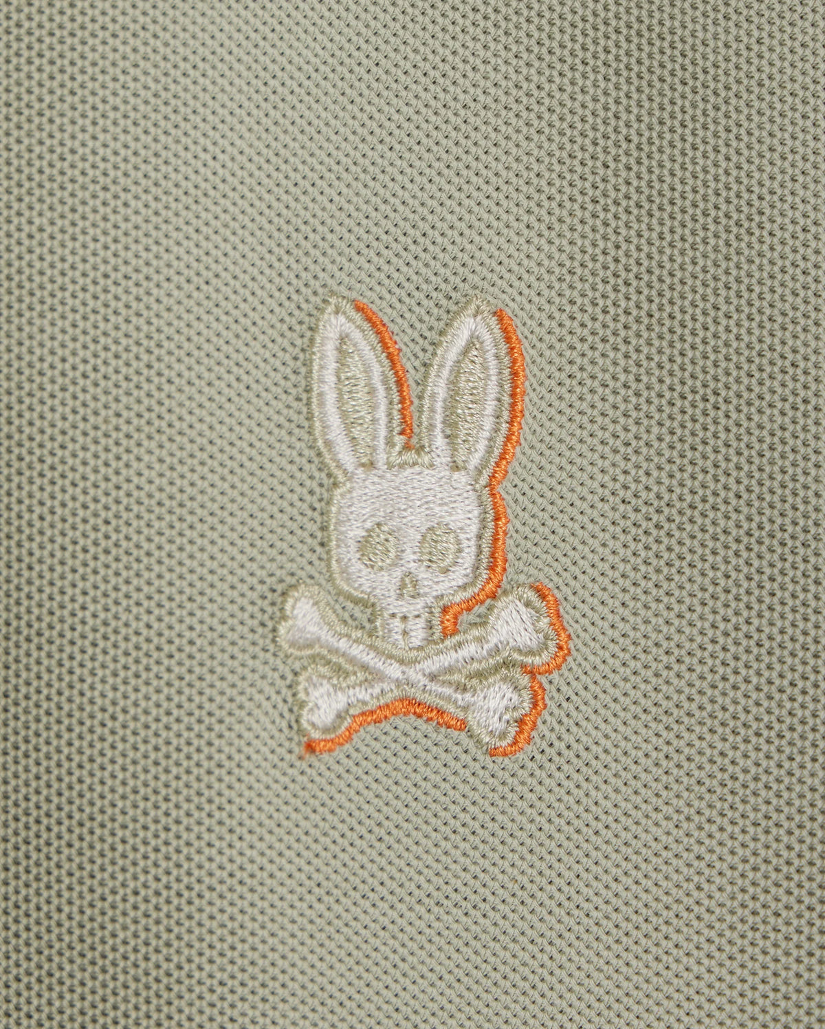 The KIDS KAYDEN PIQUE POLO (B0K675C200) by Psycho Bunny showcases a striking logo of a white embroidered bunny head with long ears, positioned above crossed bones on a light gray textured background. The bunny and bones are accentuated with orange stitching, adding an edgy flair to the classic Pima cotton pique fabric.