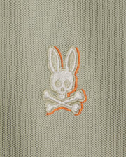 The KIDS KAYDEN PIQUE POLO (B0K675C200) by Psycho Bunny showcases a striking logo of a white embroidered bunny head with long ears, positioned above crossed bones on a light gray textured background. The bunny and bones are accentuated with orange stitching, adding an edgy flair to the classic Pima cotton pique fabric.