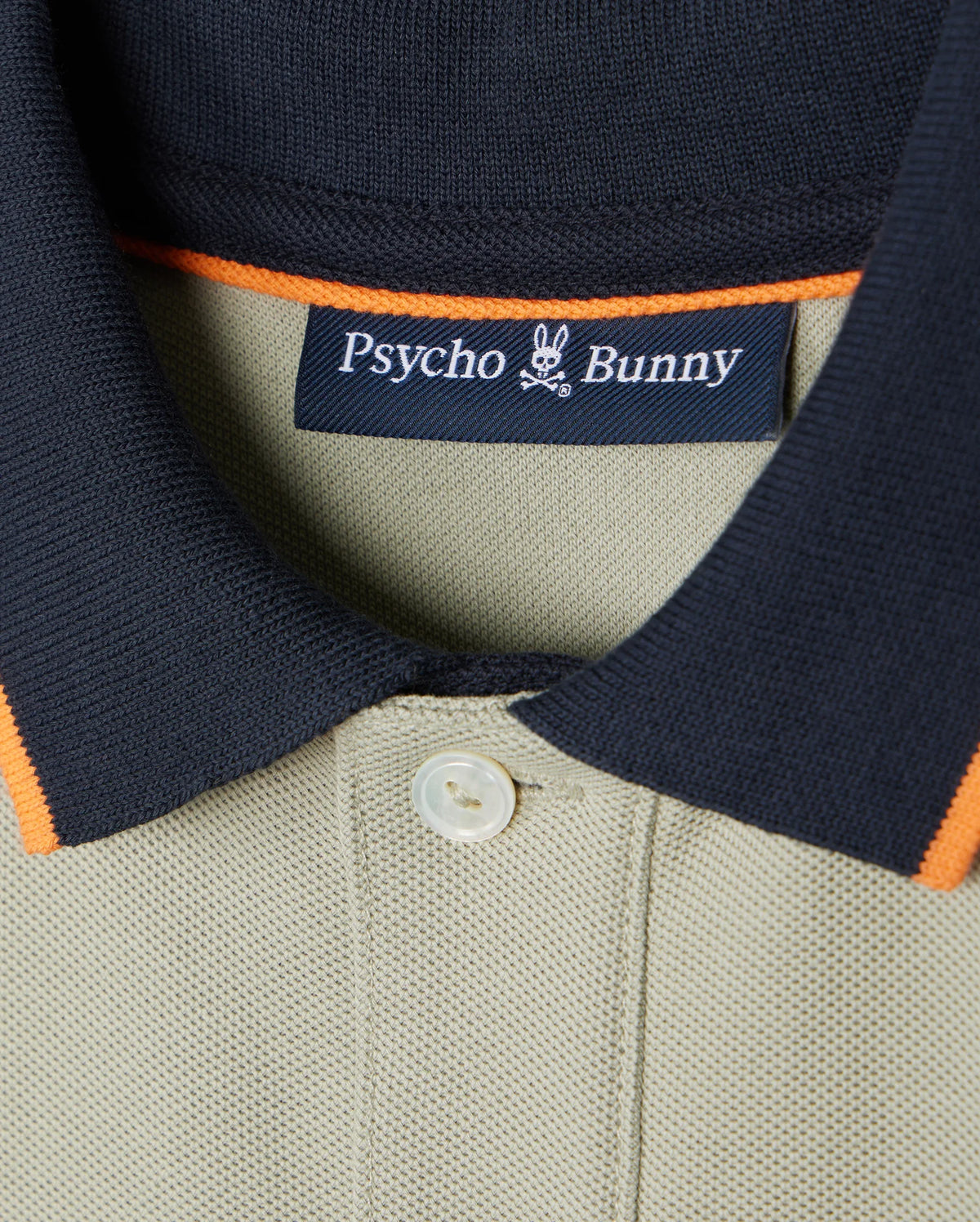 A close-up of the Kids Kayden Pique Polo - B0K675C200, featuring a khaki color with a black collar and orange trim, crafted from soft Pima cotton pique. The shirt displays the Psycho Bunny brand label inside the collar, showcasing the iconic bunny logo with crossbones below it. A single white button is visible beneath contrasting stripes on the placket.