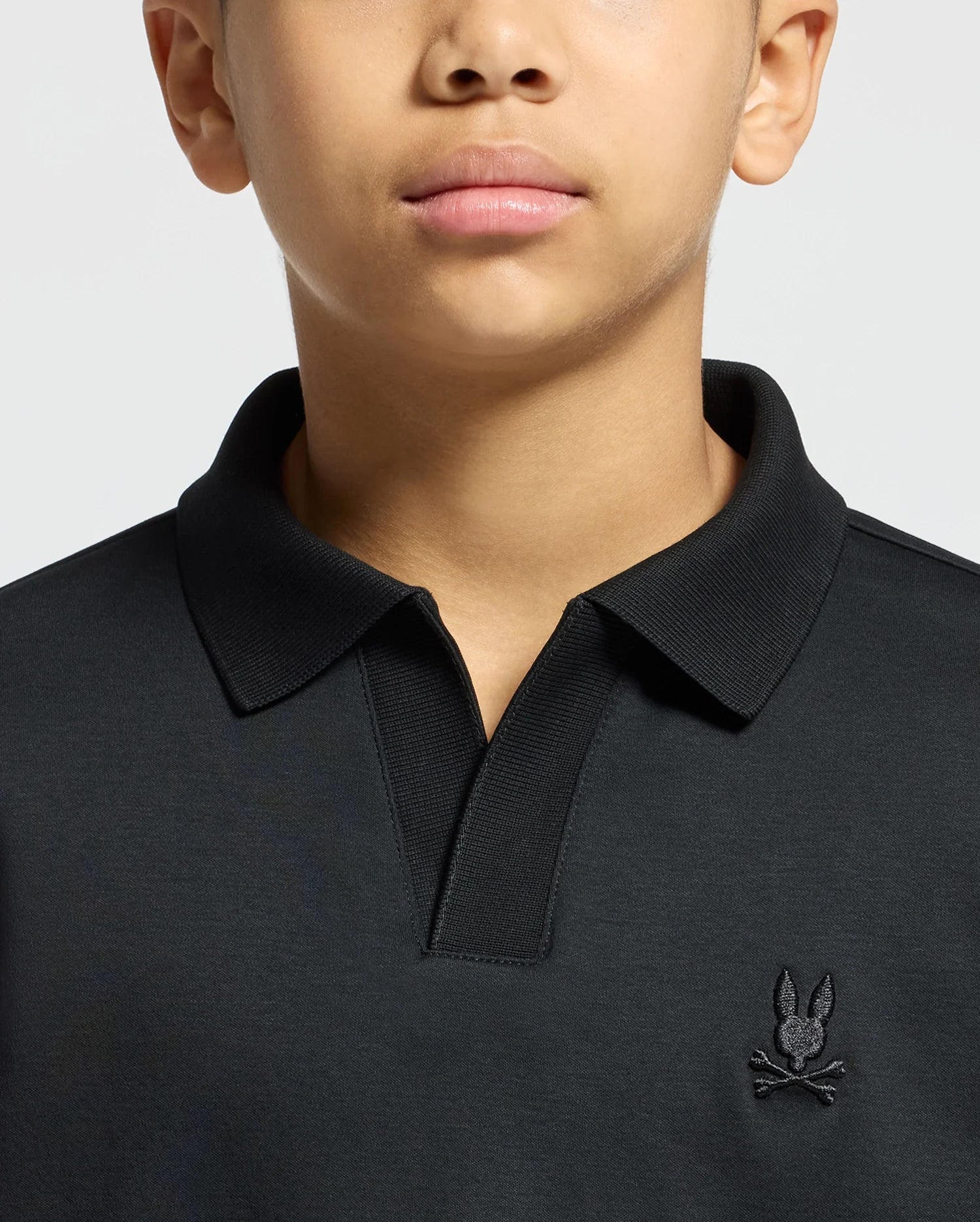 A person is wearing a black Psycho Bunny Kids Broadway Pima Interlock Johnny Collar Polo, showcasing a small embroidered rabbit with crossbones logo on the chest. The image highlights the sleek design of the upper torso and neck area.