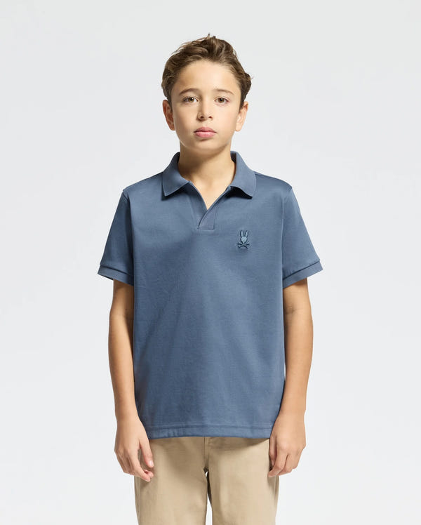 A young boy is posed against a plain background, dressed in Psycho Bunny's KIDS BROADWAY PIMA INTERLOCK JOHNNY COLLAR POLO (B0K891D200) in blue, highlighted by a small chest logo, and beige pants. The mercerized Pima cotton shirt enhances his simple yet stylish look.