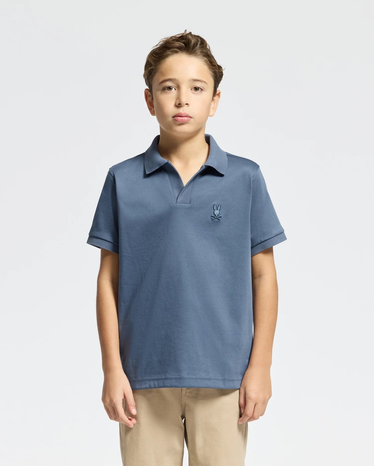 A young boy is posed against a plain background, dressed in Psycho Bunny's KIDS BROADWAY PIMA INTERLOCK JOHNNY COLLAR POLO (B0K891D200) in blue, highlighted by a small chest logo, and beige pants. The mercerized Pima cotton shirt enhances his simple yet stylish look.
