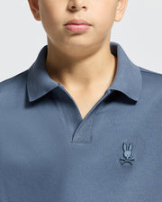 A child wearing a blue "KIDS BROADWAY PIMA INTERLOCK JOHNNY COLLAR POLO - B0K891D200" by Psycho Bunny, made from soft mercerized Pima cotton and featuring a small bunny and crossbones logo on the chest. The image highlights the upper torso and collar.