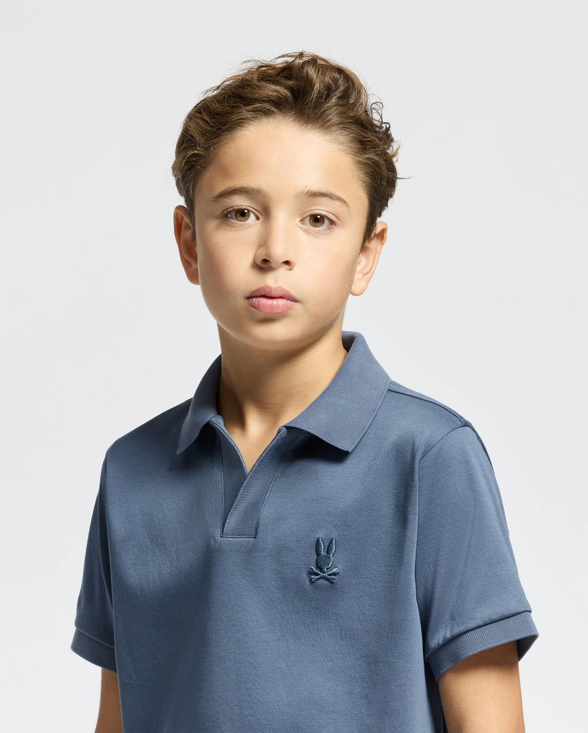 A young boy with short, light brown hair is wearing a Psycho Bunny KIDS BROADWAY PIMA INTERLOCK JOHNNY COLLAR POLO, featuring an embroidered rabbit logo. He stands against a plain, light background, looking directly at the camera.
