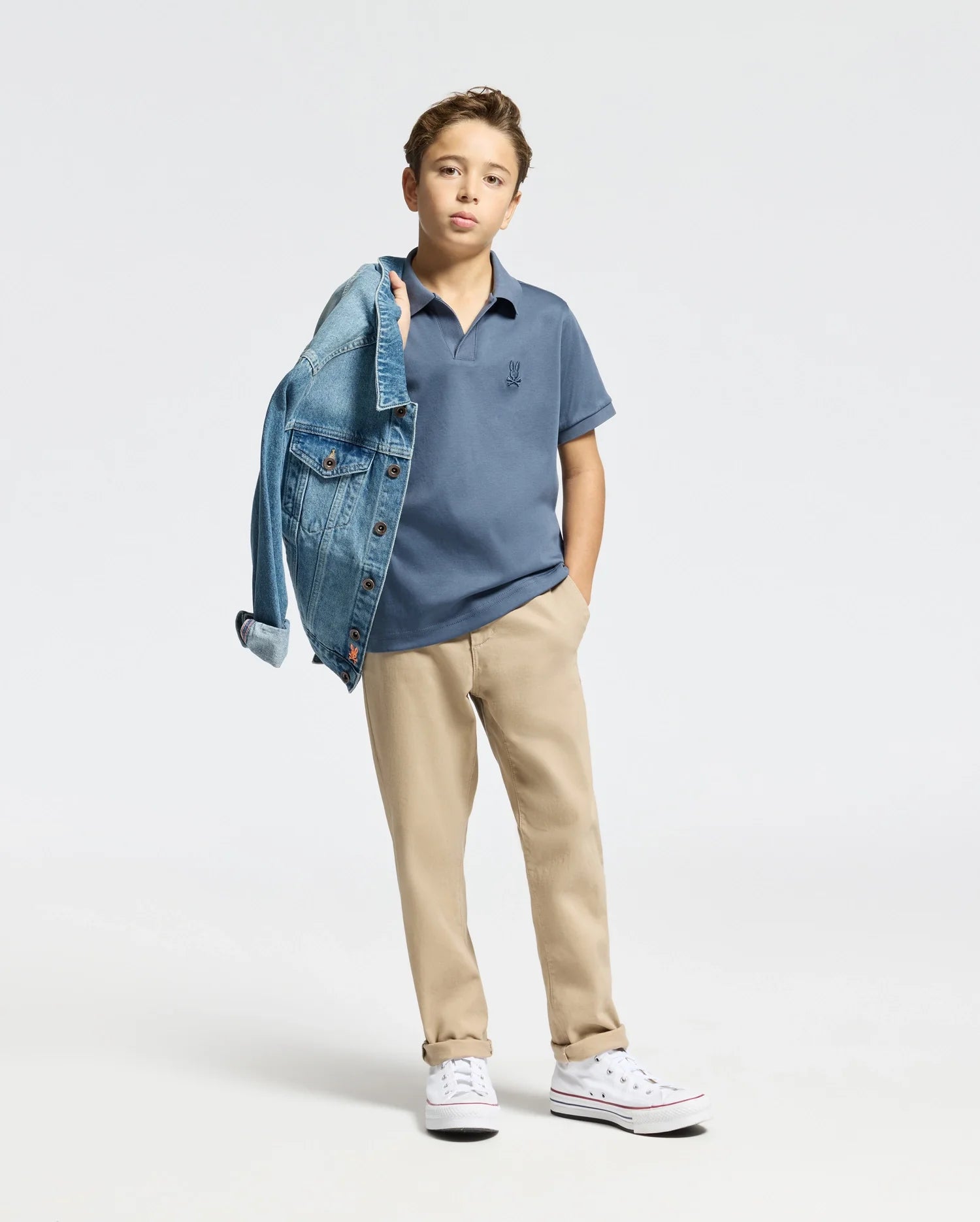 Against a plain white background, a young boy stands confidently in khaki pants and a Psycho Bunny Kids Broadway Pima Interlock Johnny Collar Polo (B0K891D200), casually draping a denim jacket over his shoulder. He completes the look with white sneakers, maintaining eye contact with the camera.