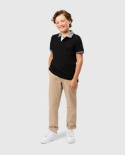 A smiling young boy with curly hair stands against a plain background. He is wearing a black KIDS MALTON PIQUE POLO SHIRT - B0K950D200 by Psycho Bunny made from Pima cotton piqué, beige trousers, and white sneakers. His hands are casually placed in his pockets.