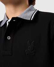 Close-up of a person wearing the Kids Malton Pique Polo Shirt B0K950D200 by Psycho Bunny, featuring a small emblem of a bunny skull and crossbones on the chest. Crafted from premium Pima cotton piqué, the shirt has a checkered collar with black and white stripes, and two buttons on the placket. The background is plain and light-colored.