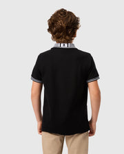 A young person with curly brown hair stands facing away from the camera, wearing a Psycho Bunny KIDS MALTON PIQUE POLO SHIRT - B0K950D200 in black with white trim on the collar and sleeves, paired with khaki pants. The background is plain light gray.