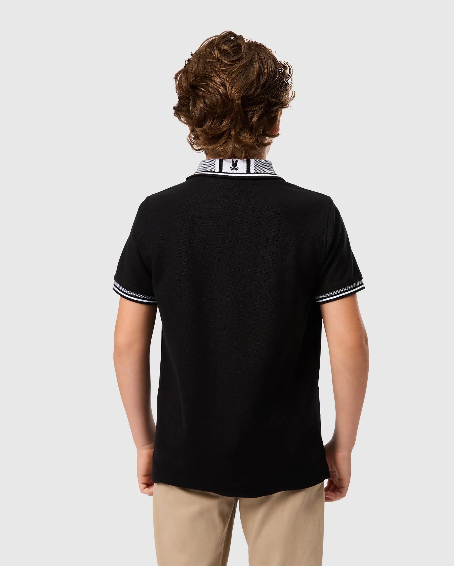 A young person with curly brown hair stands facing away from the camera, wearing a Psycho Bunny KIDS MALTON PIQUE POLO SHIRT - B0K950D200 in black with white trim on the collar and sleeves, paired with khaki pants. The background is plain light gray.