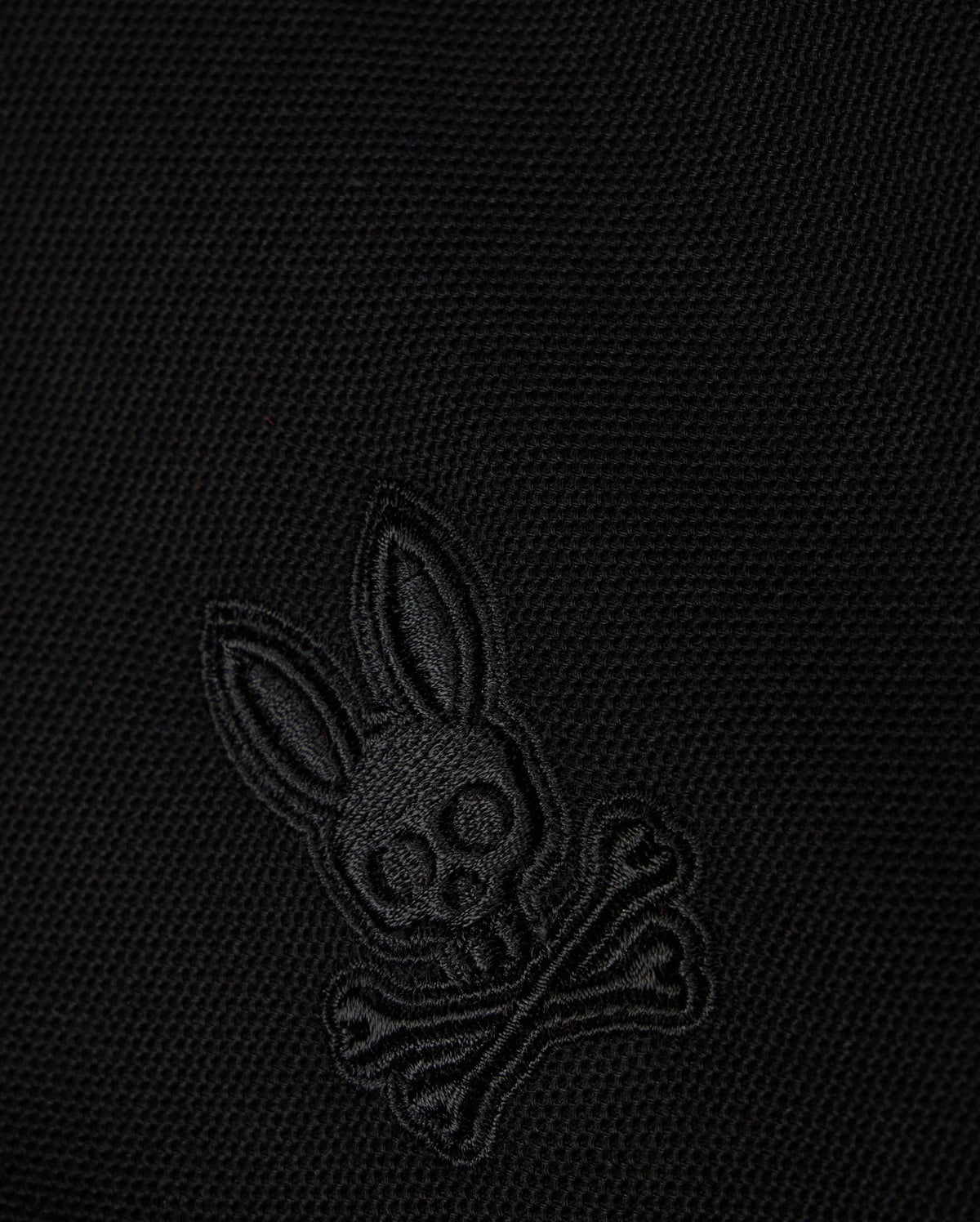 Close-up of a black Pima cotton piqué fabric with an embroidered Psycho Bunny logo on the KIDS MALTON PIQUE POLO SHIRT - B0K950D200: featuring a skull with elongated bunny ears above two crossed bones, all in a matching black color.