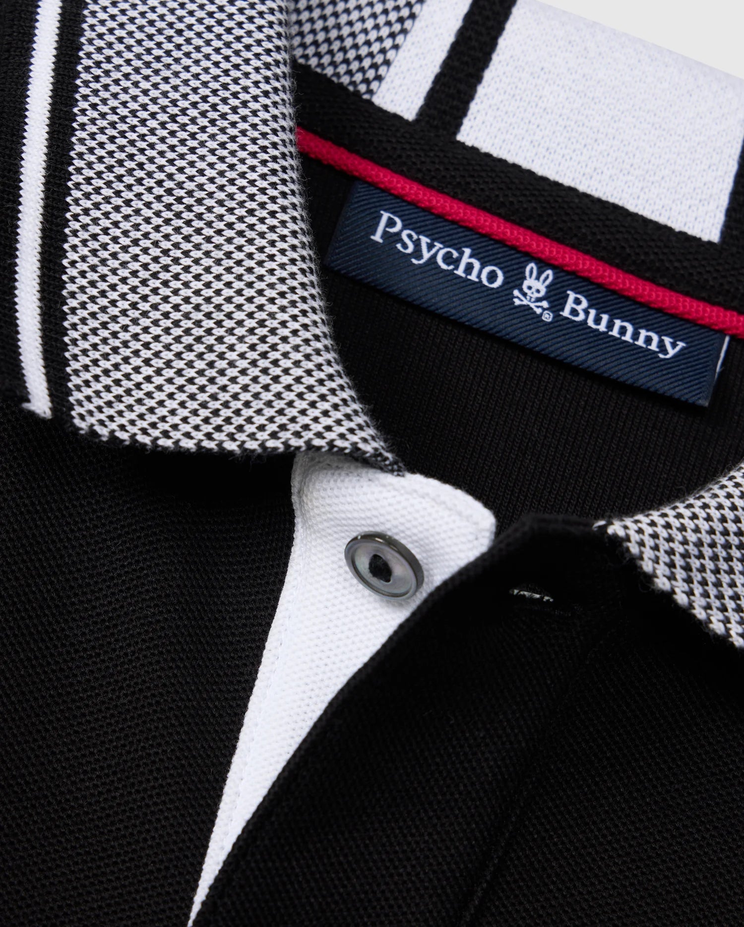 Close-up view of the KIDS MALTON PIQUE POLO SHIRT - B0K950D200 by Psycho Bunny, crafted from Pima cotton piqué. This black shirt features a distinctive black and white checkered collar with black, white, and gray stripes, complemented by a white button. The Psycho Bunny label is visible on the inside of the collar.