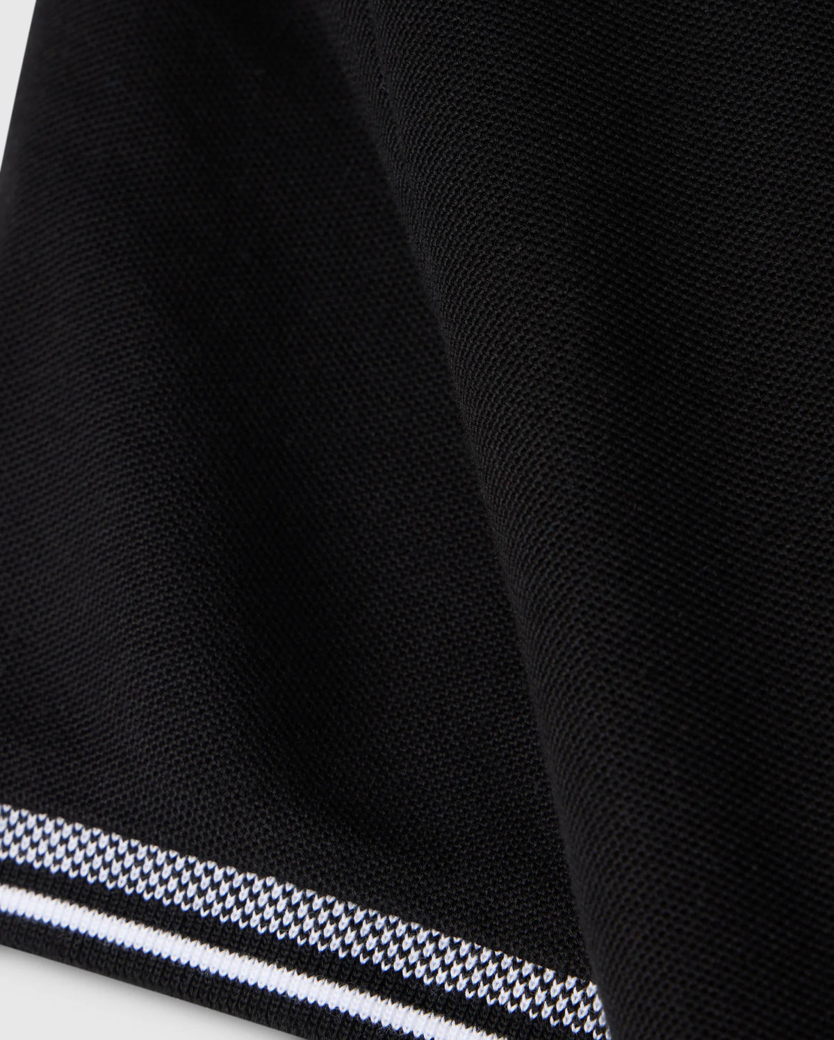 Close-up of the Psycho Bunny KIDS MALTON PIQUE POLO SHIRT - B0K950D200 in black fabric with a textured surface. The edge features a striped trim with alternating black and white lines, creating a subtle pattern contrast. The Pima cotton piqué fabric appears folded, adding a slight curve to the design, reminiscent of the Malton polo style.
