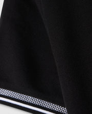 Close-up of the Psycho Bunny KIDS MALTON PIQUE POLO SHIRT - B0K950D200 in black fabric with a textured surface. The edge features a striped trim with alternating black and white lines, creating a subtle pattern contrast. The Pima cotton piqué fabric appears folded, adding a slight curve to the design, reminiscent of the Malton polo style.