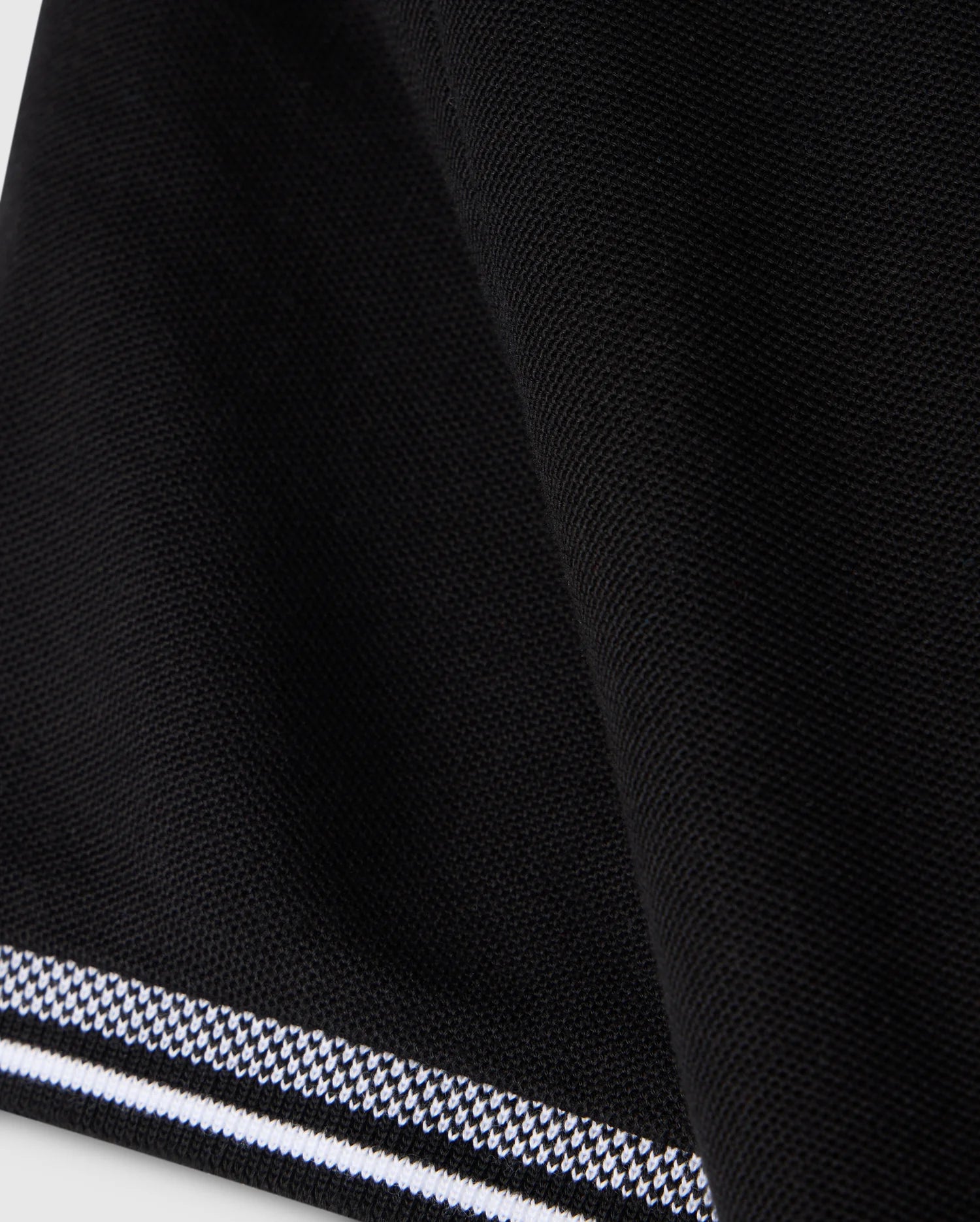 Close-up of the Psycho Bunny KIDS MALTON PIQUE POLO SHIRT - B0K950D200 in black fabric with a textured surface. The edge features a striped trim with alternating black and white lines, creating a subtle pattern contrast. The Pima cotton piqué fabric appears folded, adding a slight curve to the design, reminiscent of the Malton polo style.