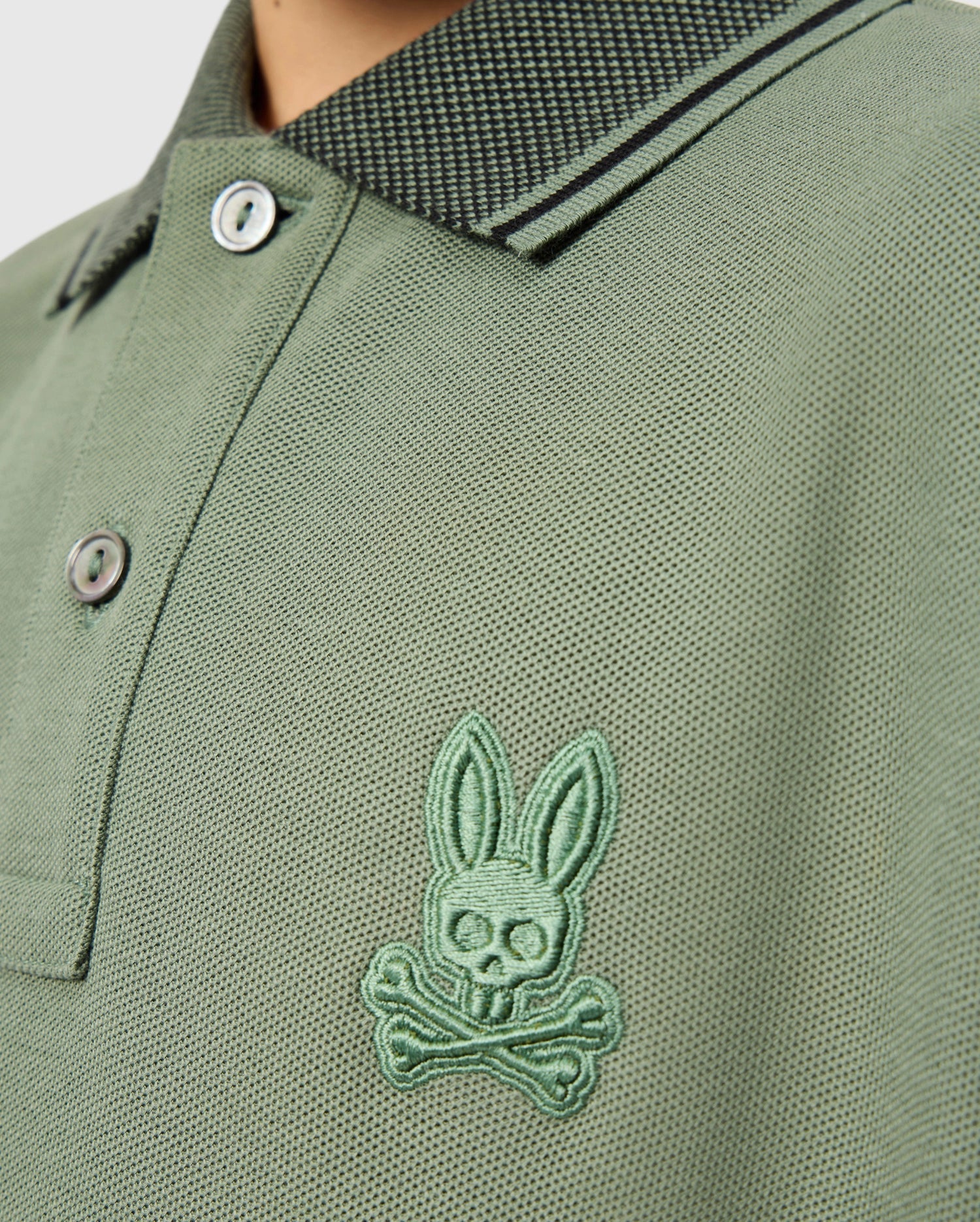 A detailed look at the KIDS MALTON PIQUE POLO SHIRT - B0K950D200 by Psycho Bunny, this green polo is made from soft pima cotton and showcases a skull-and-crossbones logo with rabbit ears on the chest. The design includes a patterned collar, diamond piqué-patterned cuffs, and two buttons for added style.