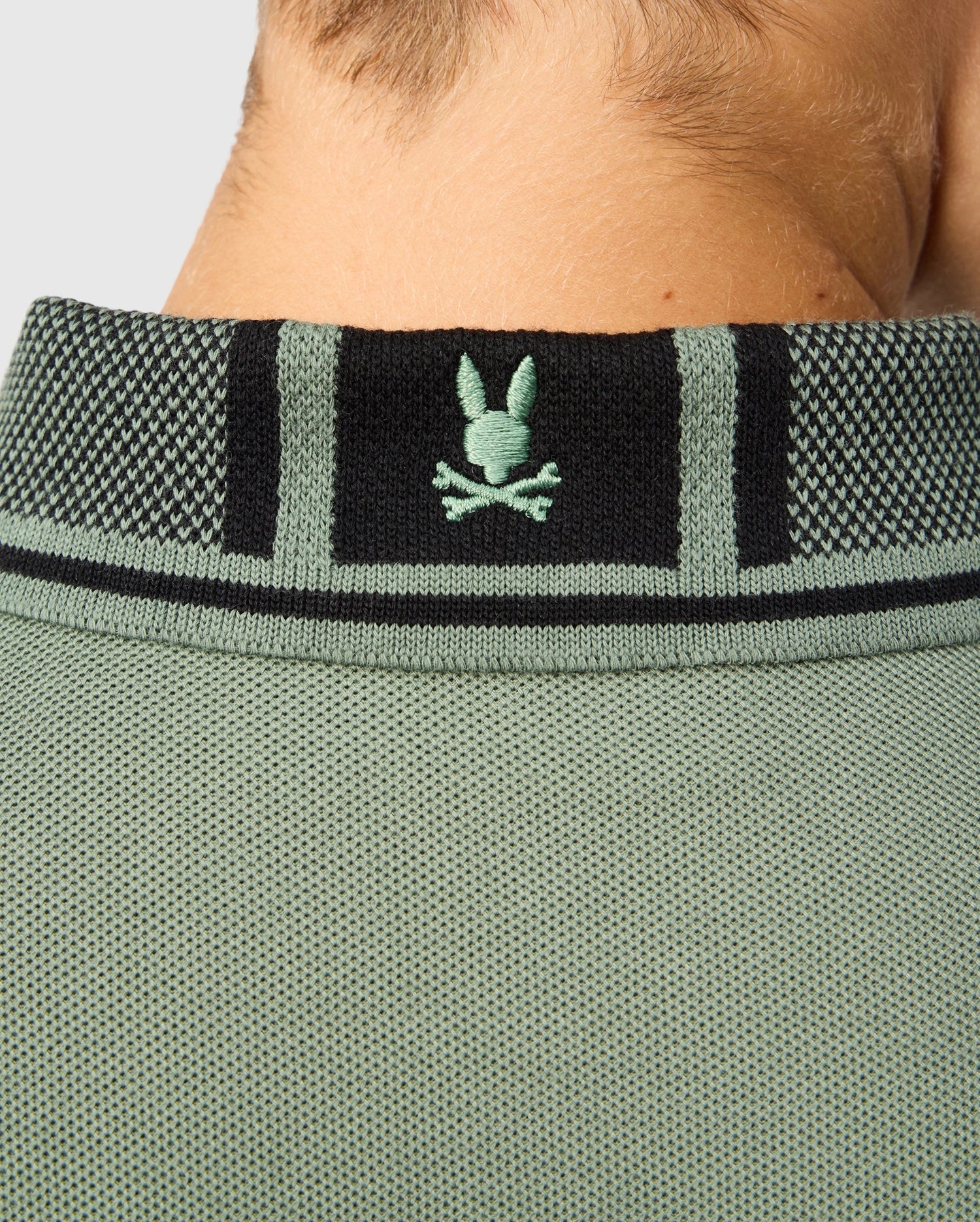 Close-up of the back of a neck wearing a green textured shirt crafted from soft pima cotton, featuring a black and green collar. The collar is adorned with a green embroidered bunny and crossbones logo, adding character to this Kids Malton Pique Polo Shirt by Psycho Bunny.