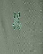 Close-up of a green Kids Malton Piqué Polo Shirt by Psycho Bunny, crafted from soft pima cotton. It showcases a small embroidered design featuring a stylized rabbit's head with crossed bones underneath. The shirt is enhanced with distinctive diamond piqué-patterned cuffs for an added touch of sophistication.