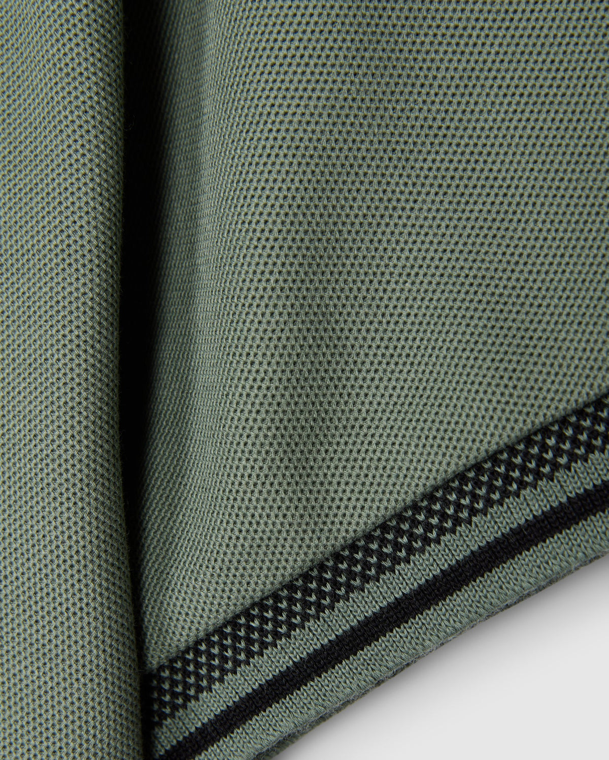 The image displays a close-up of a textured green fabric featuring small black dots and two black striped edges, similar to the style of the KIDS MALTON PIQUE POLO SHIRT by Psycho Bunny. The material, which resembles pima cotton, is draped or folded to highlight its weave and diamond piqué-patterned cuffs.