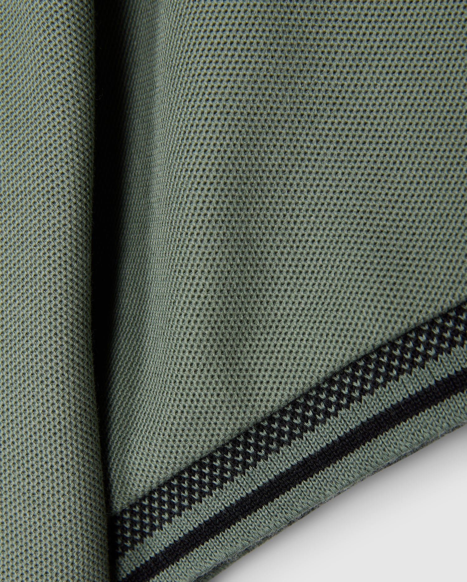 The image displays a close-up of a textured green fabric featuring small black dots and two black striped edges, similar to the style of the KIDS MALTON PIQUE POLO SHIRT by Psycho Bunny. The material, which resembles pima cotton, is draped or folded to highlight its weave and diamond piqué-patterned cuffs.