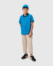 A boy is standing against a plain background, smiling at the camera. He is wearing a blue Psycho Bunny Kids Malton Pique Polo Shirt (B0K950D200), which features mother-of-pearl buttons, beige pants, black and orange sneakers, and a matching blue cap. His hands are behind his back.