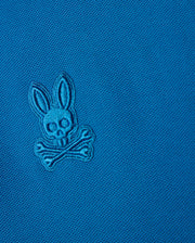Close-up of the Psycho Bunny Kids Malton Pique Polo Shirt (B0K950D200) in luxurious Pima cotton piqué, featuring an embroidered logo with a stylized skull, bunny ears, and crossbones. The design is stitched in the same blue color as the fabric, creating a subtle, tonal effect. Mother-of-pearl buttons add an elegant finishing touch.