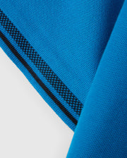 Close-up of the corner of a bright blue Pima cotton piqué fabric with a textured weave from the KIDS MALTON PIQUE POLO SHIRT - B0K950D200 by Psycho Bunny. The edge is accented with a black stripe running parallel to the fabric's border. The fabric shows a folded corner, displaying both the front and reverse sides, reminiscent of a classic Malton polo.
