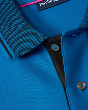 Close-up of a blue KIDS MALTON PIQUE POLO SHIRT - B0K950D200 with a textured collar featuring a black and white checkered pattern. The shirt has a button placket, and one mother-of-pearl button is visible. The brand label "Psycho Bunny" is seen on the inside of the collar, emphasizing high-quality Pima cotton piqué fabric.