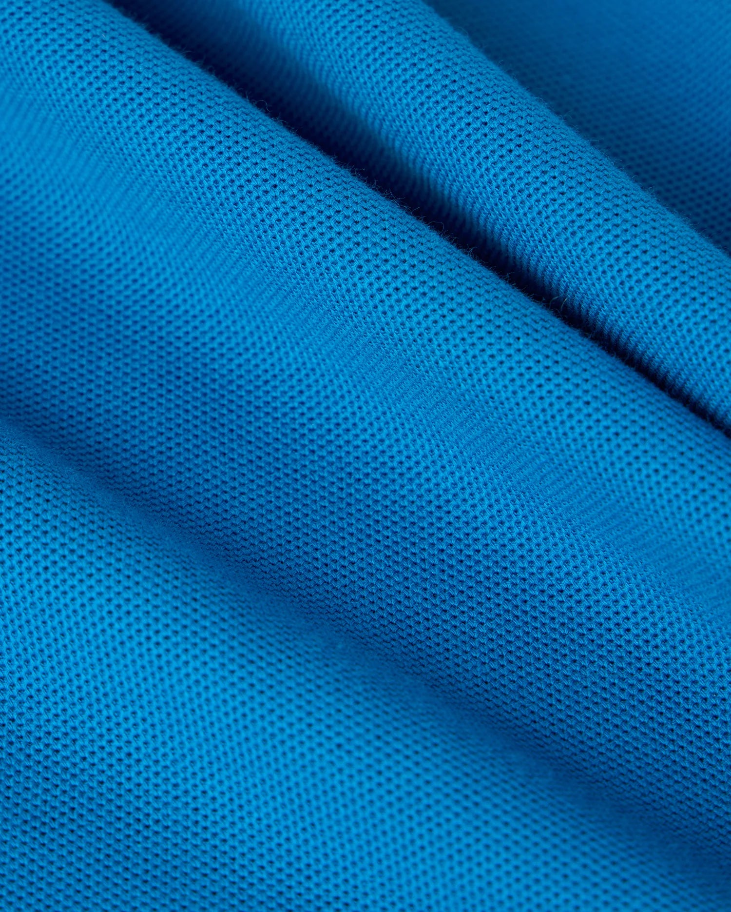 Close-up of the blue Pima cotton piqué fabric from the KIDS MALTON PIQUE POLO SHIRT - B0K950D200 by Psycho Bunny, showcasing its textured, mesh-like surface. The fabric is carefully folded, creating gentle curves and shadows that accentuate its fine, grid-like pattern. The vibrant blue color is strikingly eye-catching, harking back to the classic Malton polo shirts.