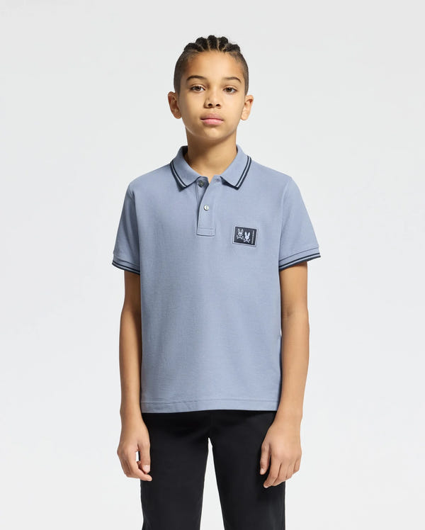 A young individual with braided hair wears the Psycho Bunny KIDS MONROE PIQUE POLO - B0K964D200, light blue in Pima cotton with a small logo appliqué, paired with black pants. The regular fit enhances their style against a plain white backdrop.