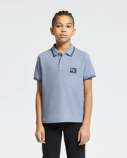 A young individual with braided hair wears the Psycho Bunny KIDS MONROE PIQUE POLO - B0K964D200, light blue in Pima cotton with a small logo appliqué, paired with black pants. The regular fit enhances their style against a plain white backdrop.