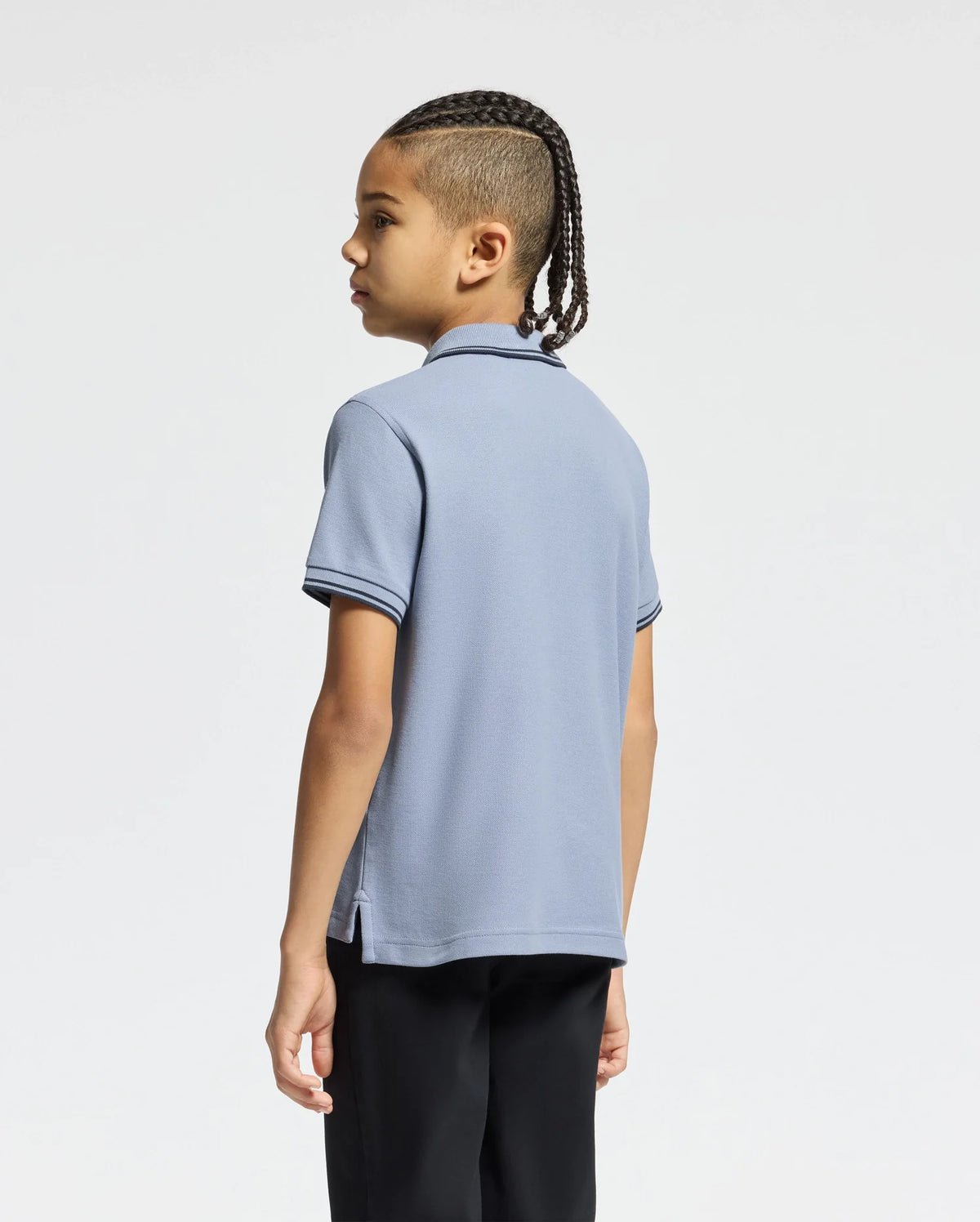 A young person with braids stands slightly left, wearing a Psycho Bunny KIDS MONROE PIQUE POLO in light blue with dark trimmings and black pants, perfectly complementing the plain white background.