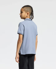A young person with braids stands slightly left, wearing a Psycho Bunny KIDS MONROE PIQUE POLO in light blue with dark trimmings and black pants, perfectly complementing the plain white background.