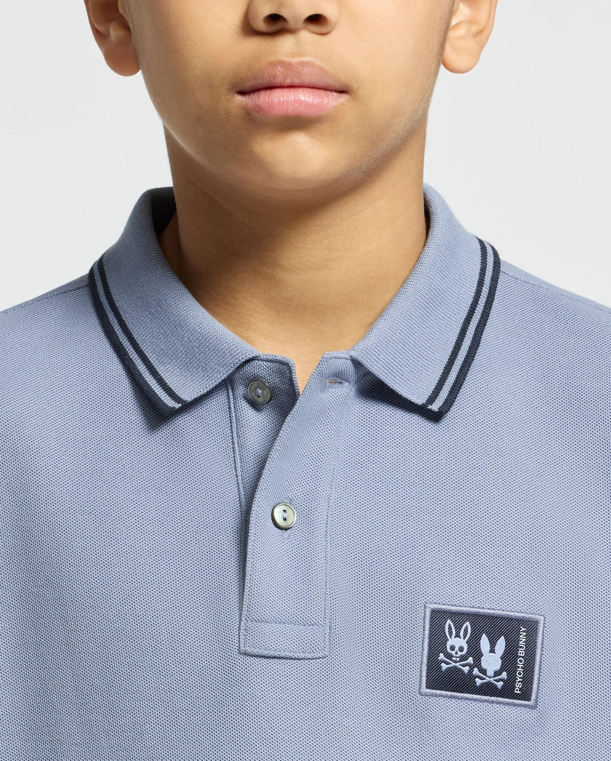 A person wearing a Psycho Bunny KIDS MONROE PIQUE POLO in light blue, showcasing a bunny with crossed bones logo appliqué on the chest and dark collar trim. The regular fit conceals most of the face except for the chin and mouth.