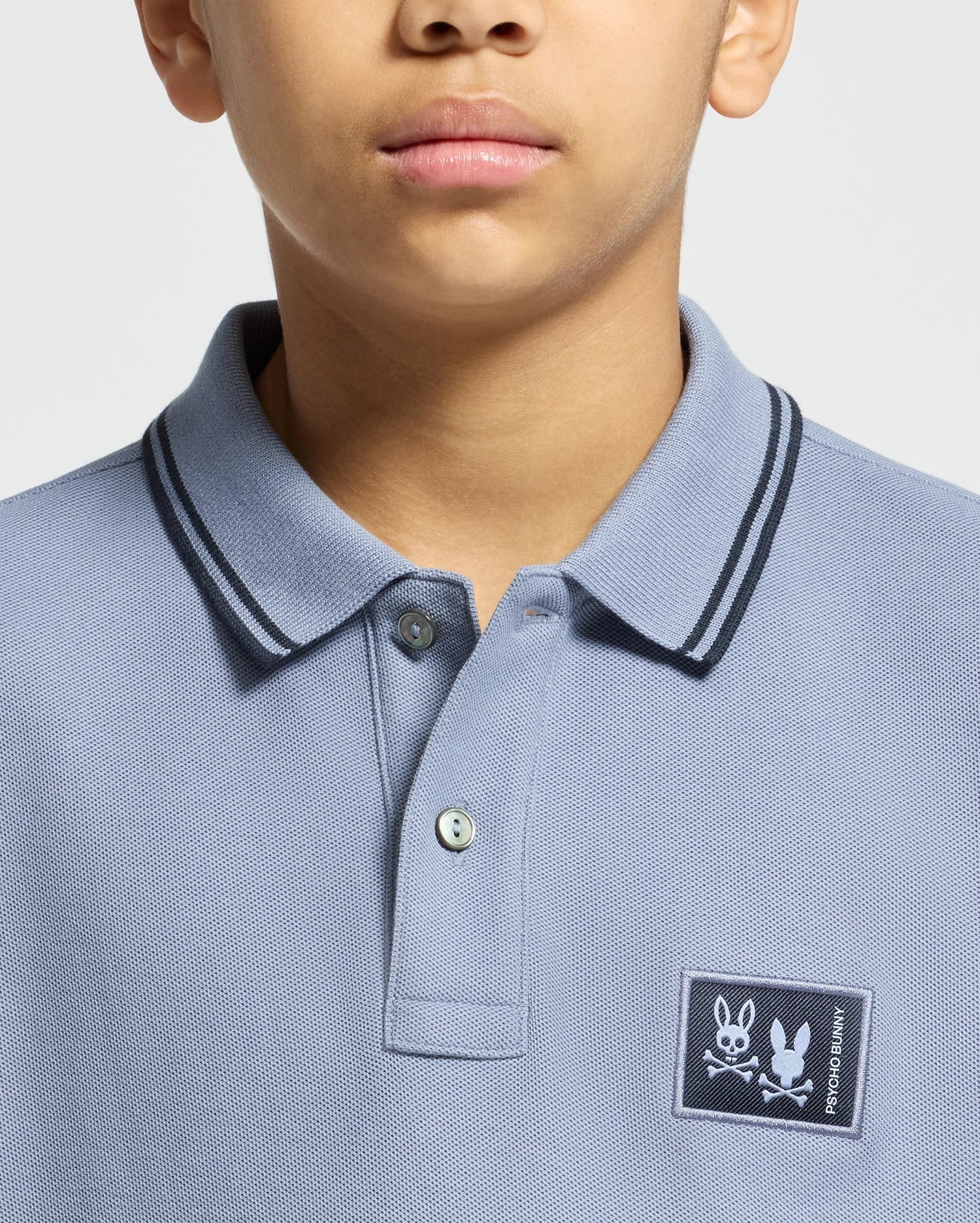 A person wearing a Psycho Bunny KIDS MONROE PIQUE POLO in light blue, showcasing a bunny with crossed bones logo appliqué on the chest and dark collar trim. The regular fit conceals most of the face except for the chin and mouth.