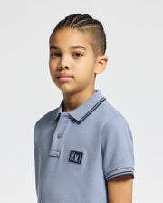 A young person with braided hair wears the Psycho Bunny KIDS MONROE PIQUE POLO in light blue, featuring a small logo appliqué. They stand against a plain white background, gazing directly at the camera with a neutral expression.