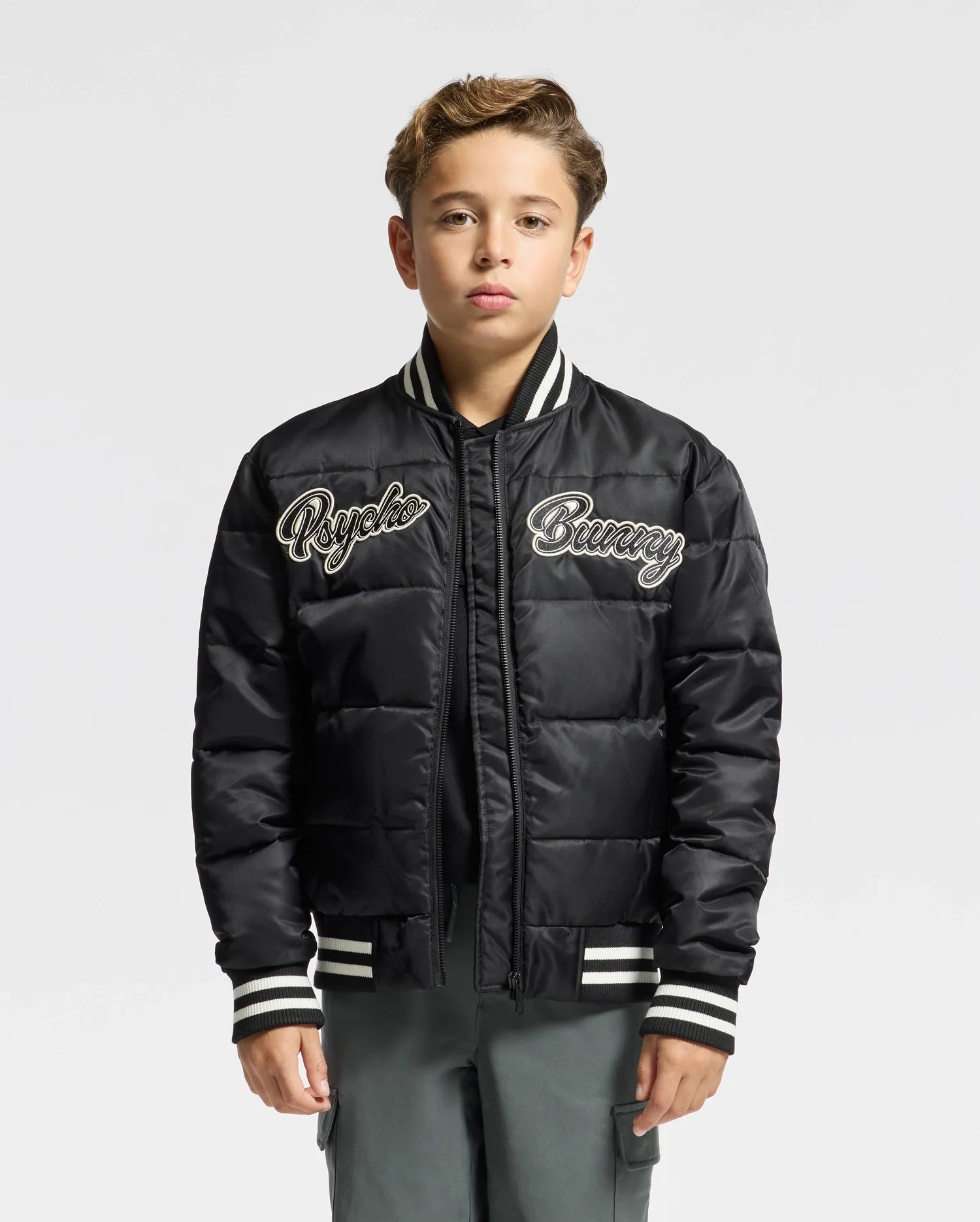 A young individual is wearing the fashionable "KIDS FRASER FASHION PUFFER - B0N338E200" by Psycho Bunny, featuring white and black striped cuffs and collar. The stylish black puffer jacket, embroidered with "Psycho Bunny," is a standout in kids' fashion, perfectly complemented by their short hair against a plain white background.